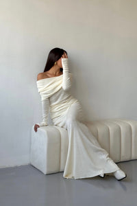 Woman in an off-shoulder, long-sleeved, ivory gown sits on a minimalist white bench against a plain background. She has long, dark hair, partially covering her face while her right arm rests on the bench and left hand touches her face.