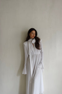 A woman with long, wavy hair wearing the Evey dress stands against a textured white wall. The flowing white dress features long sleeves, flared cuffs, and a high neck adorned with a white flower. Perfect for special occasions, she gazes downward with a serene expression.