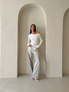 A woman stands against a light-colored wall with arched recesses, wearing the Léa top—a chic off-the-shoulder white knit piece—and silver wide-legged pants. She has long straight hair and wears white heeled sandals, with one hand casually in her pocket. The minimalist aesthetic of the setting enhances her sophisticated style.