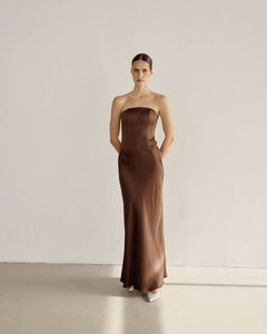 A person is standing against a plain white wall, dressed in a long, strapless brown dress made from lightweight fabric. Their hands rest in the pockets of the Silky Maxi Skirt, and they have a calm expression. The floor is light gray, and the lighting is soft.