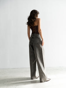 A woman with long, wavy hair stands with her back to the camera, wearing the dark, strappy Imari Corset top and high-waisted, wide-leg gray jeans—timeless essentials for any wardrobe. The background is plain and white, with a light source casting soft shadows.