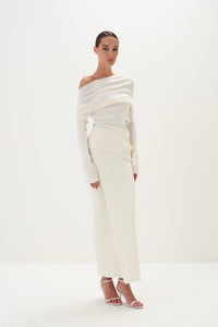 A model wearing a white asymmetric draped neckline top and a matching long skirt, with accessories and shoes.