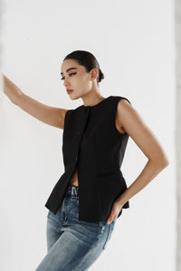 A person with dark hair tied back is standing against a white background, wearing the sleeveless button-up Eloise Vest and blue jeans. Their pose is relaxed, with one arm resting against a wall or surface. The overall impression is casual yet stylish, embodying a timeless design.