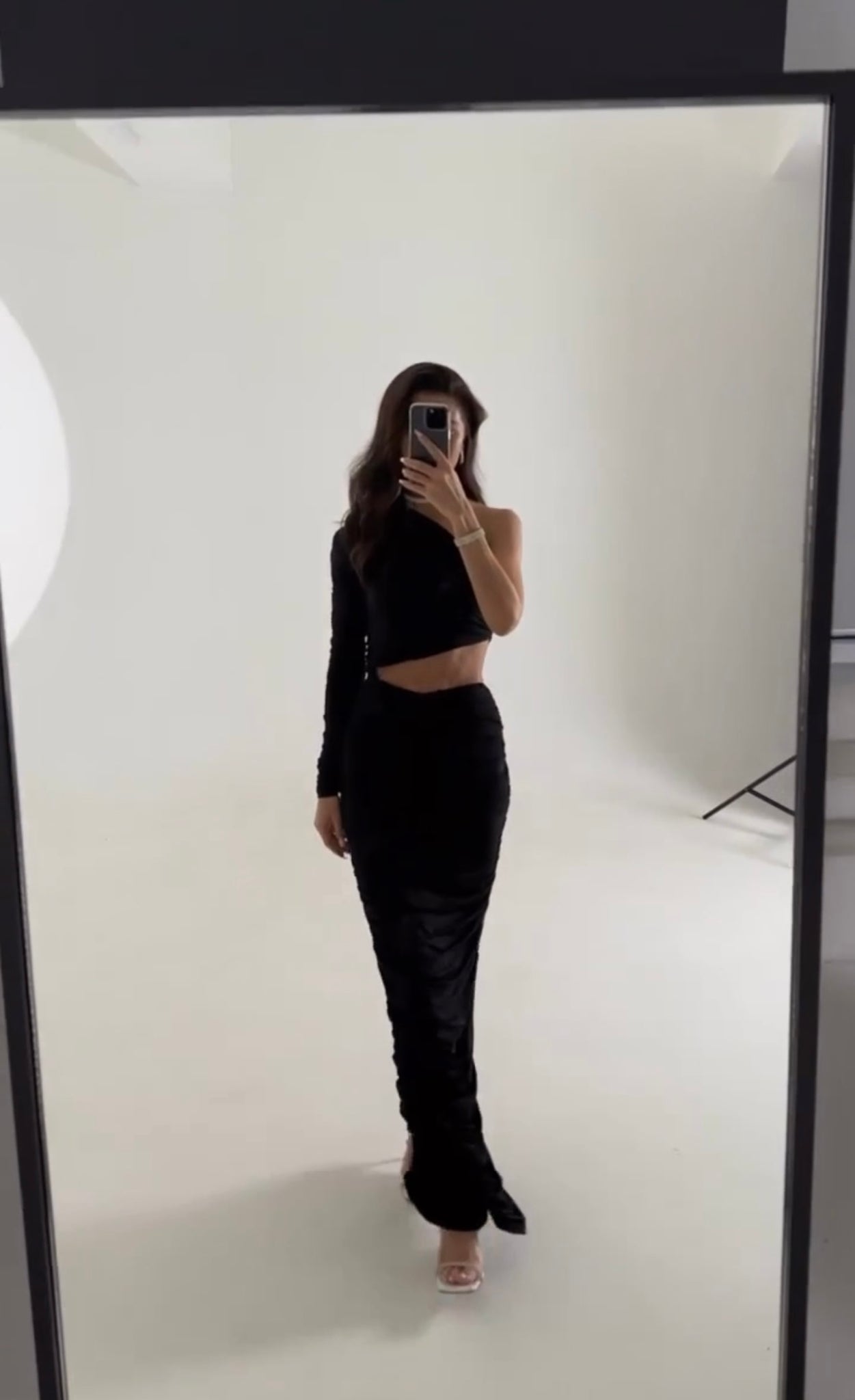 A person with long hair takes a mirror selfie in a well-lit studio, wearing a stylish, fitted black Amicia dress maxi dress featuring one long sleeve and a side cutout at the waist. They stand with one foot slightly forward, and their face is obscured by their phone.