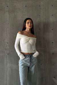 A woman with long dark hair is standing against a textured grey wall. She is wearing the Sabella top, a high-quality, soft cotton, long-sleeved, off-the-shoulder white top with ruched detailing at the chest, paired with light blue high-waisted jeans. She has a neutral expression and her hands tucked in her pockets.