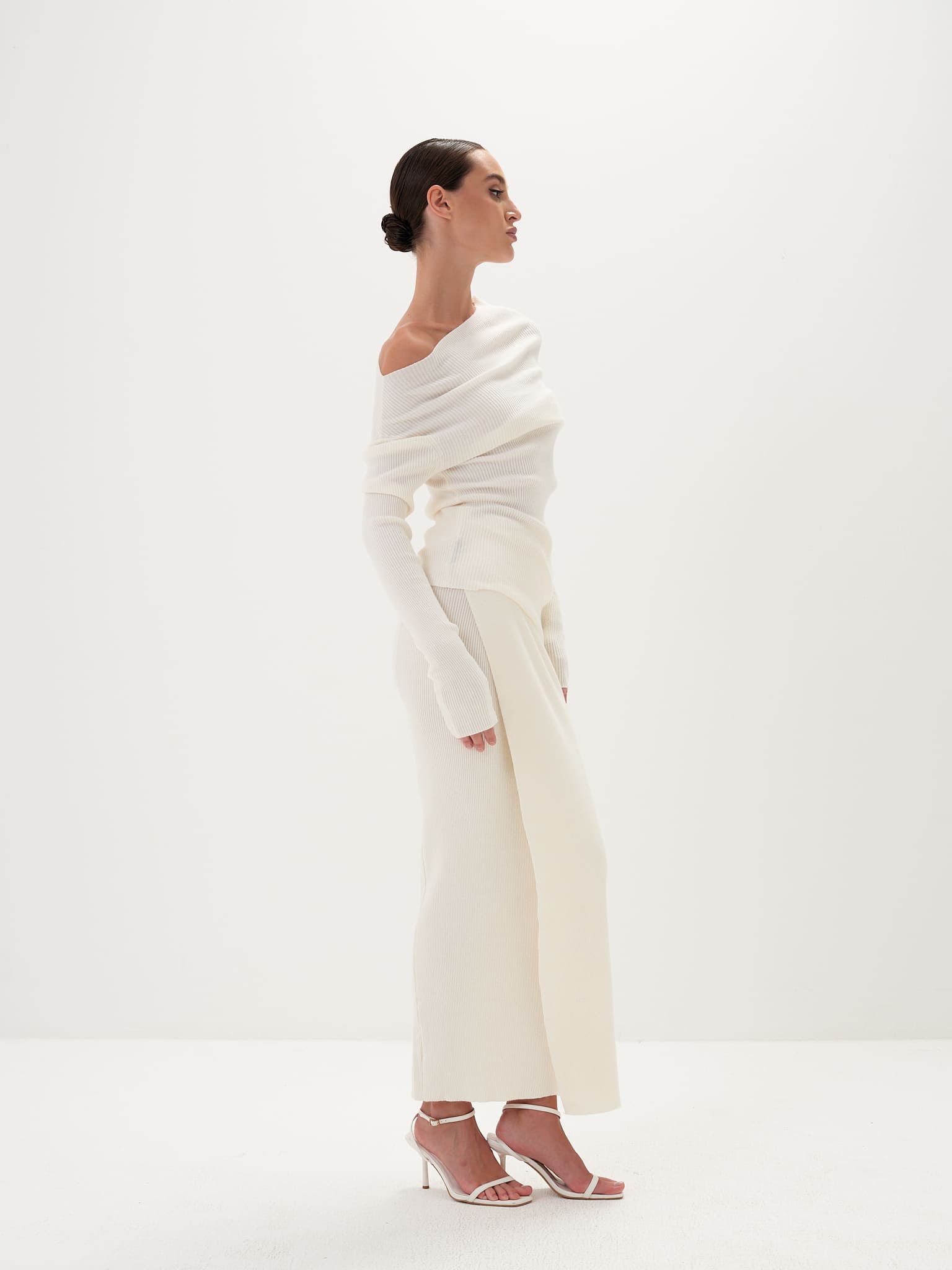 A model wearing a white asymmetric draped neckline top and a matching long skirt, with accessories and shoes.