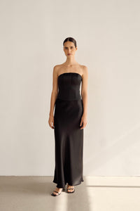 A woman stands against a plain white wall wearing the Regina Corset Set in Black, showcasing a strapless design with satin fabric, a fitted bodice, and an elegant long skirt. She complements her outfit with black open-toe sandals while her slicked-back hair adds sophistication. The lighting casts soft shadows on the wall and floor.