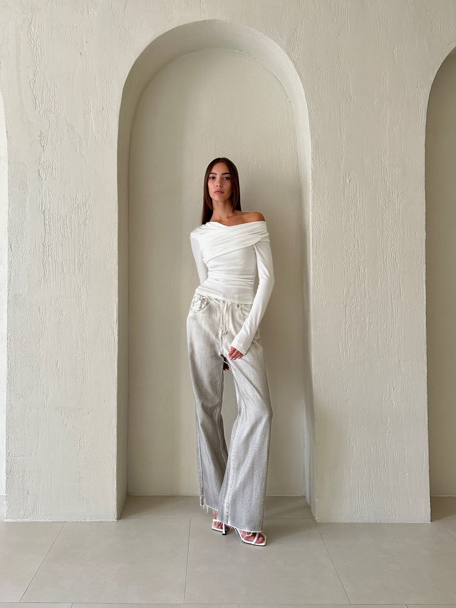 A person stands against a textured, light-colored, arched alcove. They are wearing the Léa top in an off-shoulder white knit design, paired with light gray wide-leg pants and white sandals. The outfit exudes sophisticated style and minimalism, beautifully complementing the neutral background.