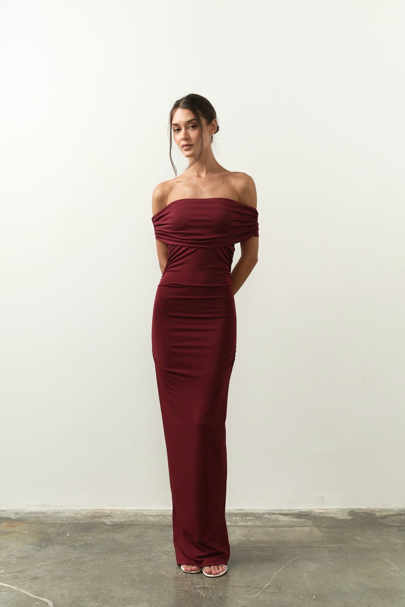 Dressed in the Scarlett Dress, a woman exudes elegance in an off-shoulder, floor-length burgundy ensemble that creates a feminine silhouette against a plain white backdrop. Her hair is effortlessly styled up, complementing her look along with her strappy sandals. With hands gracefully clasped behind her back, she gazes forward with poised confidence.