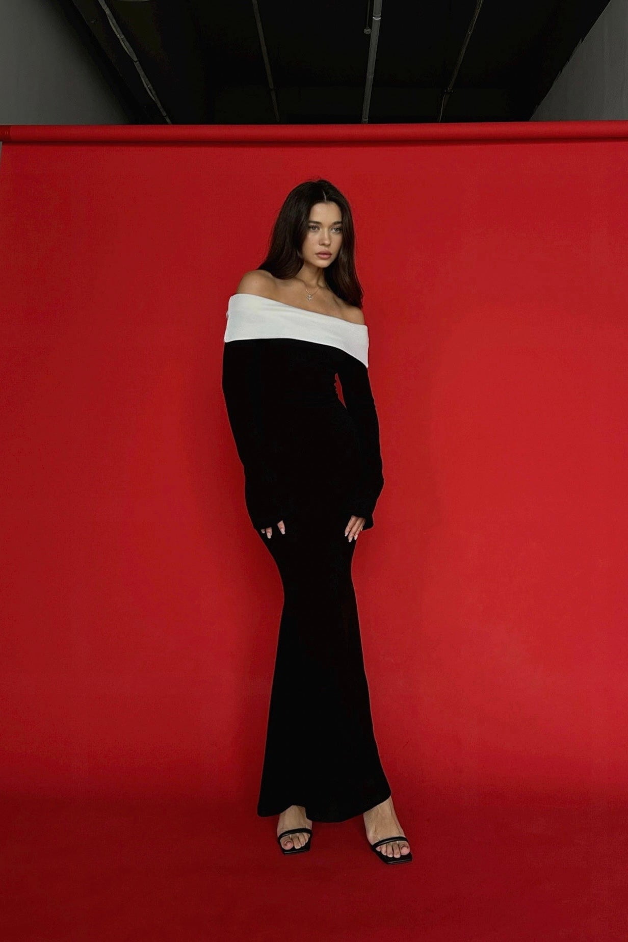 A woman stands against a red background, wearing an elegant Esber knit dress with an off-the-shoulder design and a white band at the top. She is looking at the camera with her hands by her sides and is wearing black heels.