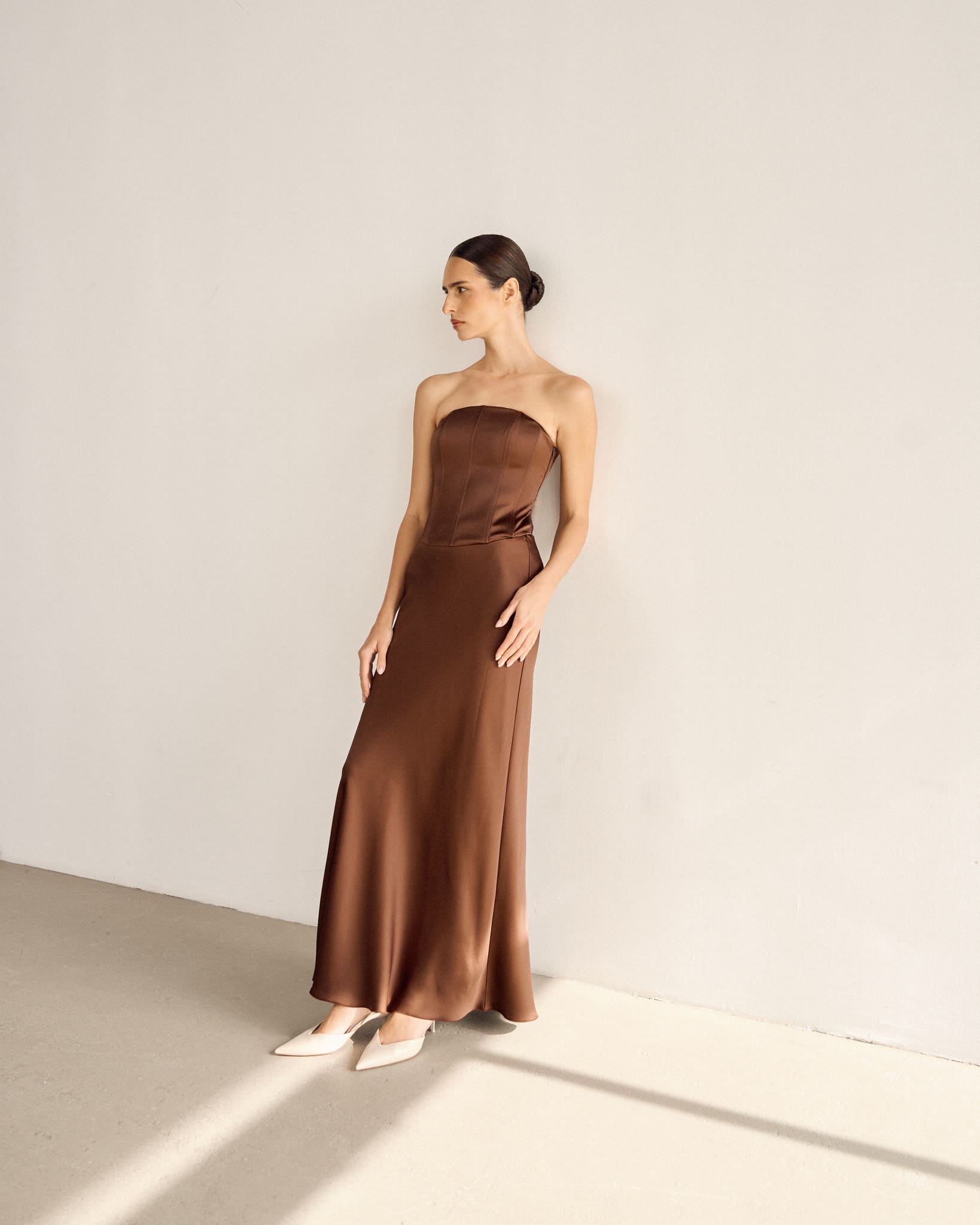 A woman in a strapless brown satin Regina corset set with a maxi skirt stands against a plain white wall. Her hair is pulled back as she gazes to her left, paired with white pointed-toe heels, while soft lighting casts subtle shadows on the floor.