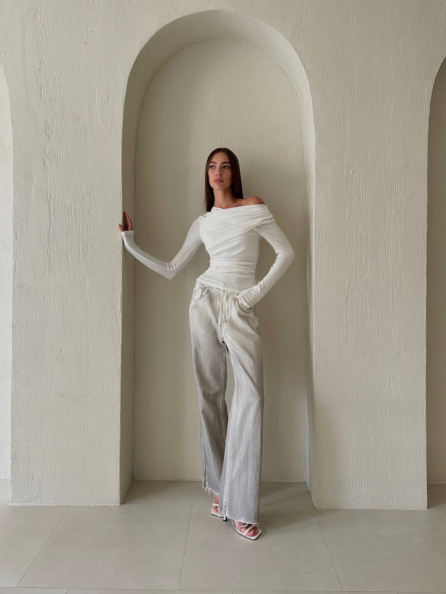 A person with long dark hair stands against an arched, textured wall. They are wearing the sophisticated Léa white knit top with long sleeves, light gray wide-leg pants made from high-quality cotton fabric, and white heeled sandals. One hand is resting on the wall while the other hangs at their side.