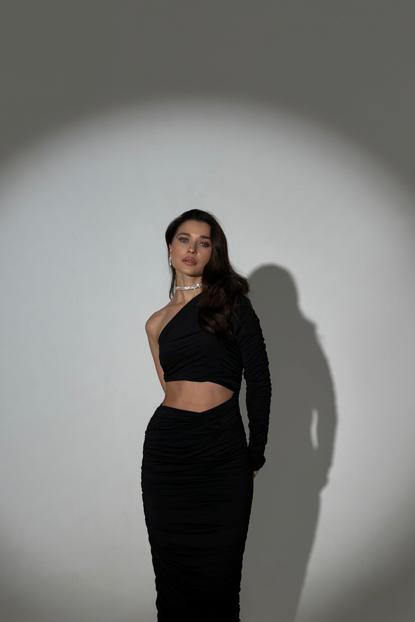 A woman stands confidently against a gray backdrop, illuminated by a spotlight that casts her shadow on the wall. She wears an elegant Amicia dress featuring a stylish one-shoulder cutout at the waist and a subtle ruched design. Her long, dark hair cascades over her shoulder.