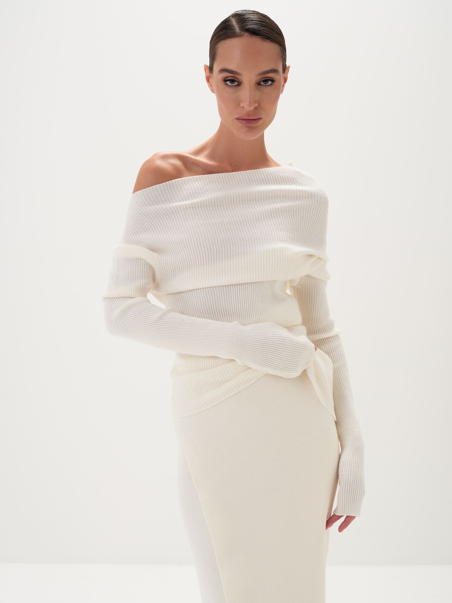 A model wearing a white asymmetric draped neckline top and a matching long skirt, with accessories and shoes.