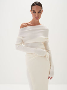 A model wearing a white asymmetric draped neckline top and a matching long skirt, with accessories and shoes.