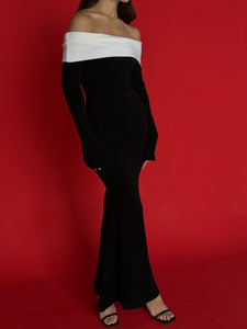 A person wearing the Esber knit dress, a black, long-sleeve, off-the-shoulder ensemble with white trim, stands against a red background. They complement the look with black open-toe heels. The body-hugging fit of the dress enhances its allure, though their head is not visible in the image.