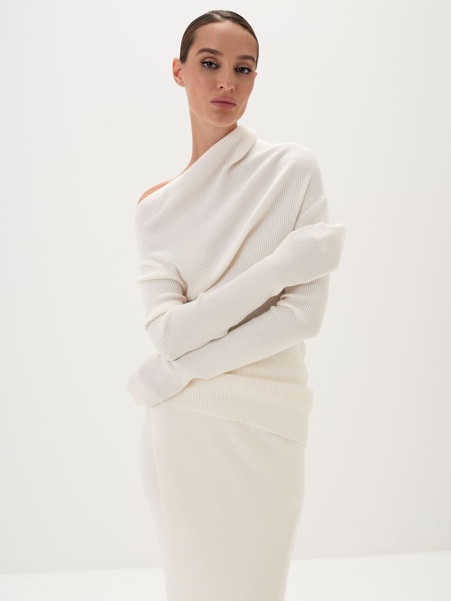 A model wearing a white asymmetric draped neckline top and a matching long skirt, with accessories and shoes.