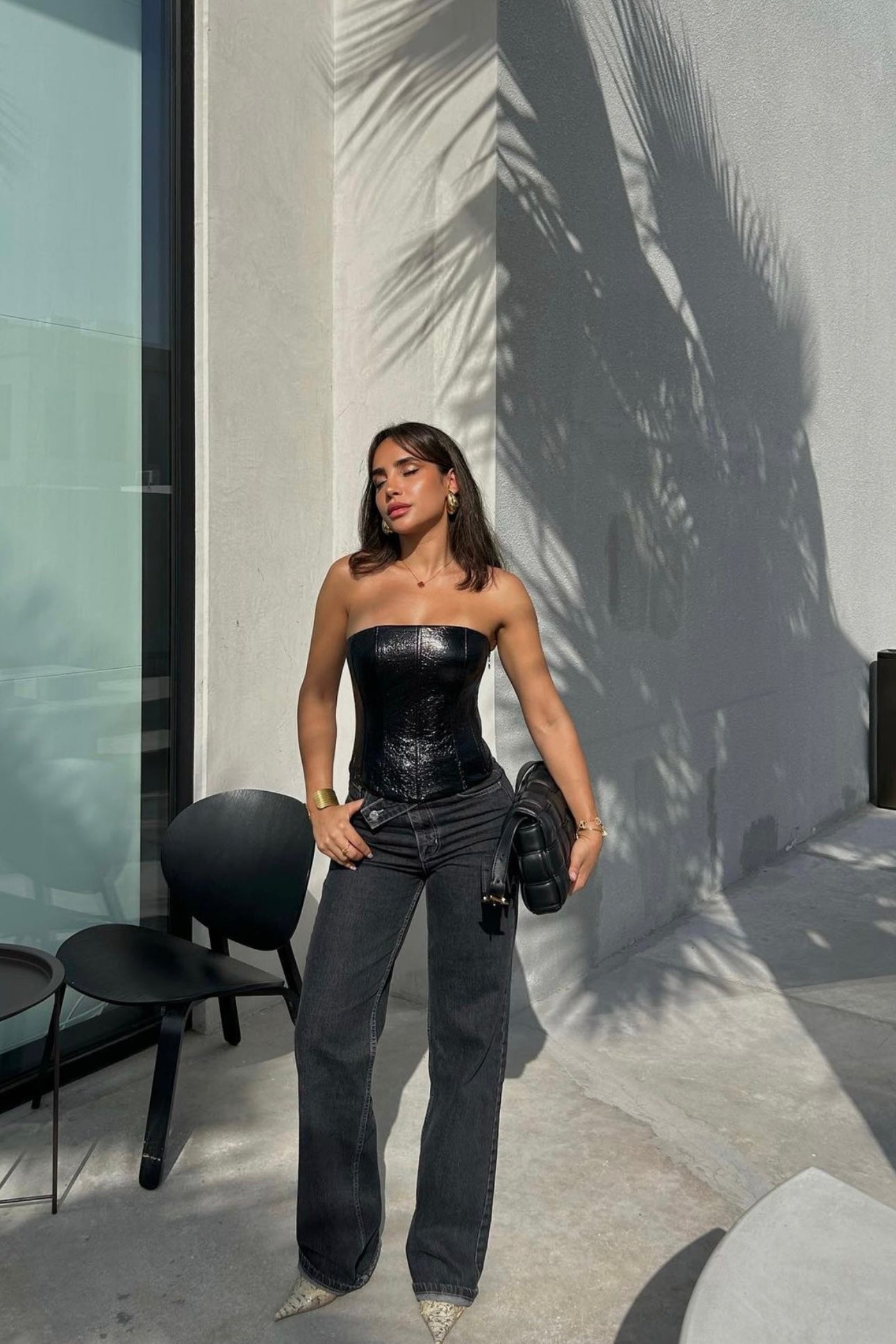 A woman stands outdoors in front of a concrete wall shadowed by palm leaves. She wears an Imari Corset, high-waisted jeans, and pointed shoes. She carries a black handbag over her left shoulder and stands next to a black chair and table. The look is a timeless essential for any wardrobe.