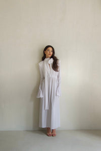 A woman stands against a plain light-colored wall, wearing the elegant Evey dress with flared sleeves and a large flower detail on the collar. Her dark hair is styled in loose waves, and she is looking calmly ahead, embodying statement fashion perfect for special occasions.