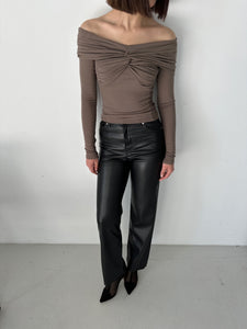 A woman stands facing forward against a plain white background, dressed in the Sabella top—a high-quality, off-the-shoulder, long-sleeved taupe top made of soft cotton fabric with a twisted front detail. She is also wearing black leather pants and black pointed-toe shoes. Only her torso and legs are visible; her head is cropped out of the frame.