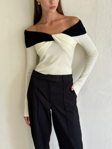 An individual exuding a sophisticated look stands against a textured white wall, donning the Amélie knit top in black and white with an off-the-shoulder twist design paired with high-waisted black pants. The person has long, straight hair and their right hand is casually placed in their pants pocket.