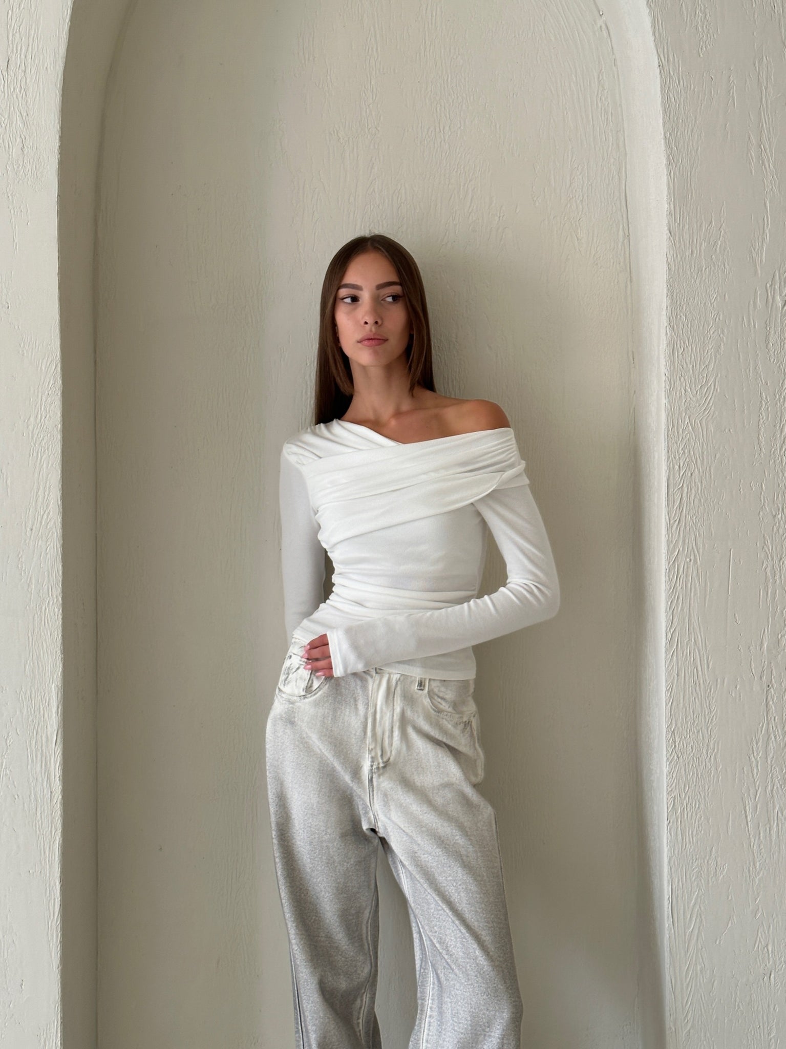 A woman stands against an arched white wall. She has long brown hair and wears the sophisticated Léa top, a white knit with long sleeves, paired with light-colored pants. Her hands are in her pockets as she gazes slightly to her left.