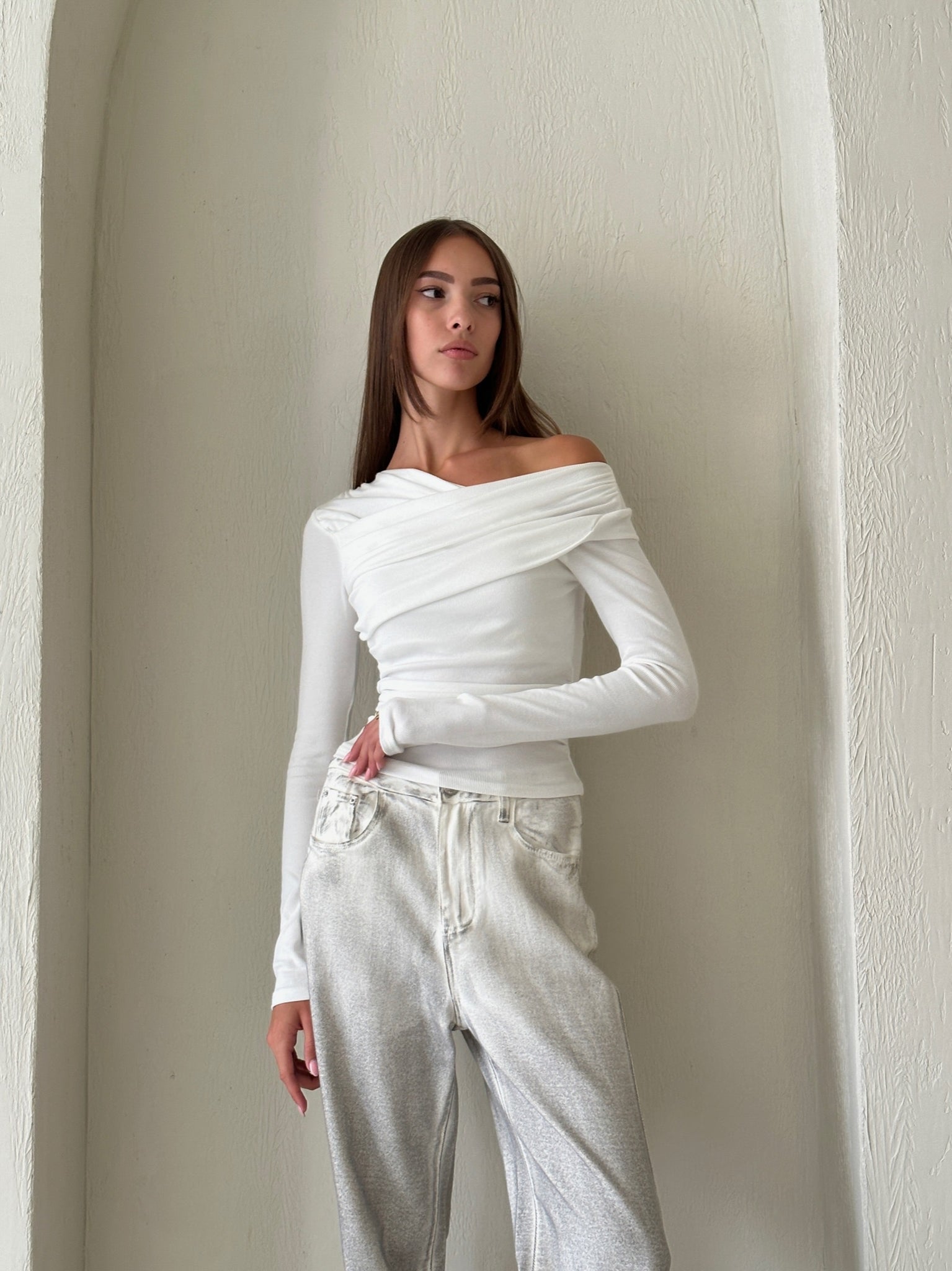 A young woman with long straight hair stands in front of a textured, arched wall. She is wearing the Léa top, an asymmetrical white off-the-shoulder piece made from high quality cotton fabric, paired with light-colored pants. Her left hand rests on her hip as she gazes to the right with a neutral expression.