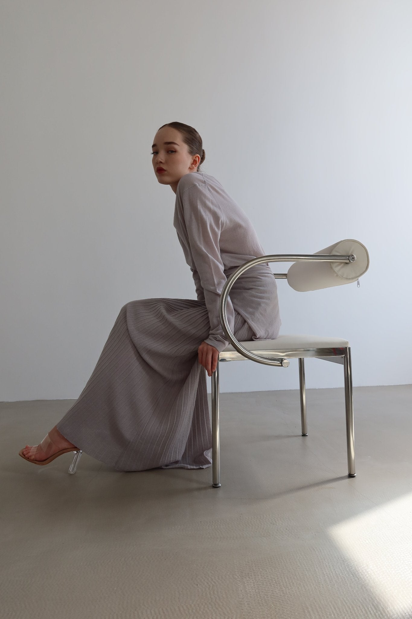 A person with sleek slicked-back hair sits sideways on a contemporary white and chrome chair in a minimalist setting. Clad in the Alaya Set, which showcases butter-soft fabric with a long-sleeved top and light grey ruffled skirt, they gaze thoughtfully into the distance.