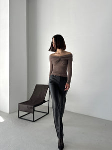 A person with a bob haircut is standing in a minimalist room. They are wearing the Sabella top, an off-the-shoulder, long-sleeve garment crafted from high-quality soft cotton fabric, along with high-waisted pants. A sleek, modern chair is placed against the white wall behind them, casting a soft shadow on the floor.