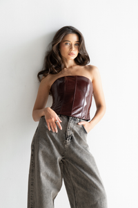 Woman with long dark hair wearing a strapless, shiny maroon Imari Corset and loose-fitting grey jeans, posing confidently against a plain white background. This timeless essential accentuates her style perfectly.