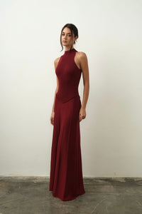 A woman wearing the Asteri Dress, featuring a sleeveless halter neck design in maroon, stands against a plain white wall. Her hair is styled in a loose updo as she gazes forward with a neutral expression, while the concrete floor introduces contrasting texture to the image.