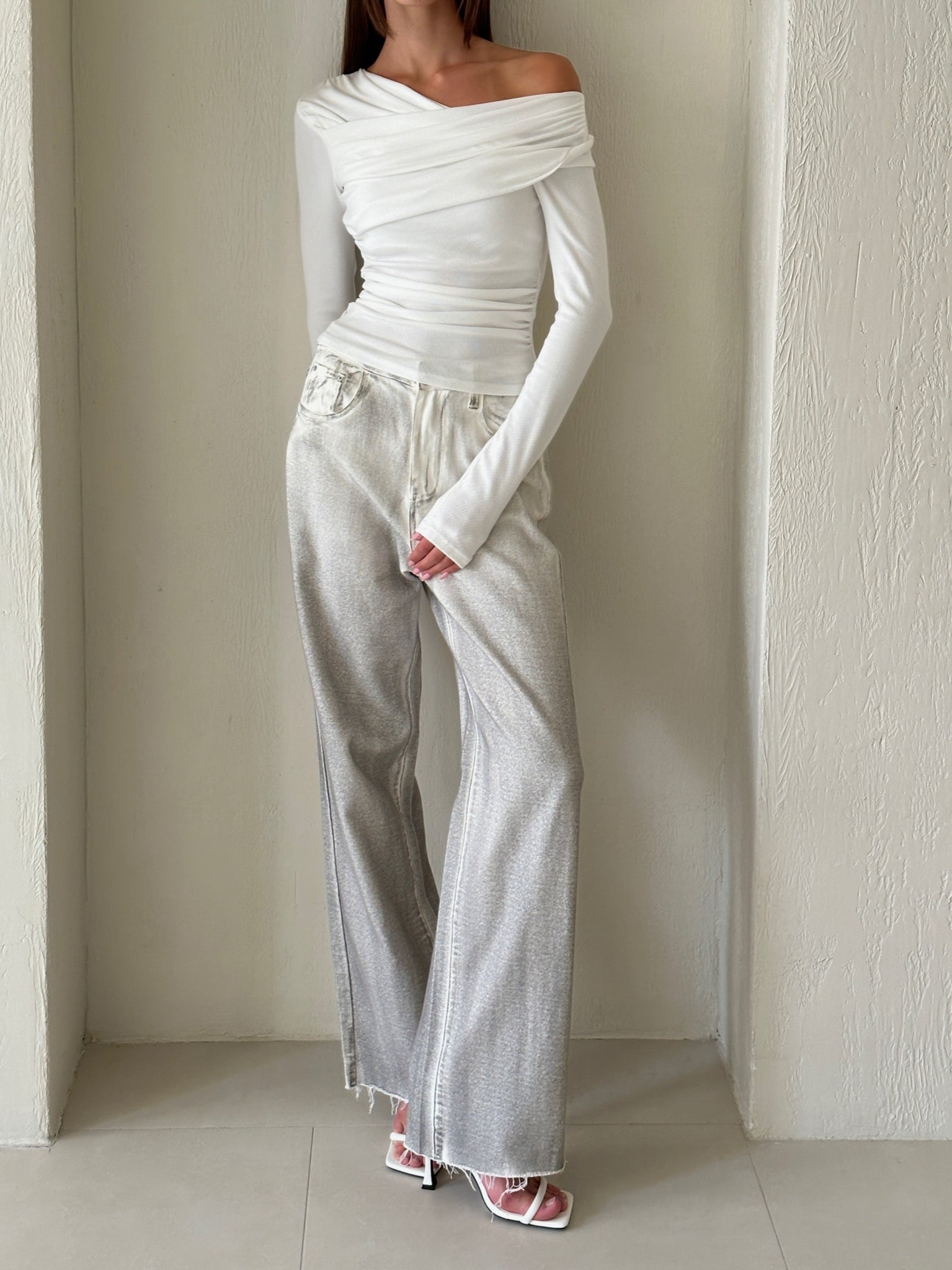 Against a textured white wall, an individual showcases a chic outfit featuring the fitted, off-shoulder Léa top with long sleeves and high-waisted, loose-fitting light-colored pants. With one hand casually placed in their pocket and paired with white heeled sandals, their partially visible hair complements the sophisticated style.