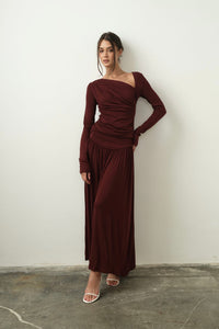 A woman stands against a plain wall, elegantly dressed in the Ariana Set, showcasing a burgundy off-shoulder top with long sleeves paired with a matching full-length skirt. She poses confidently with one hand on her hip and her hair styled up, complemented by chic white sandals.