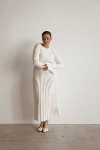 A person stands against a minimal, uncluttered white backdrop, donning the Abel Dress—a long, white garment featuring a pleated texture and figure-skimming silhouette. They have short hair pulled back and are posed with arms crossed, looking down slightly.