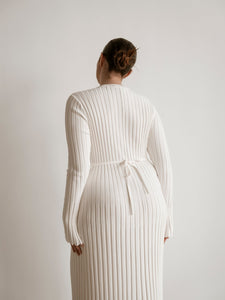 A person with their hair in a bun is standing and facing away from the camera. They are wearing an Abel Dress, which is a long, light-colored dress with a figure-skimming silhouette and long sleeves. The dress features a tie at the waist and pleated texture. The background is plain and white.