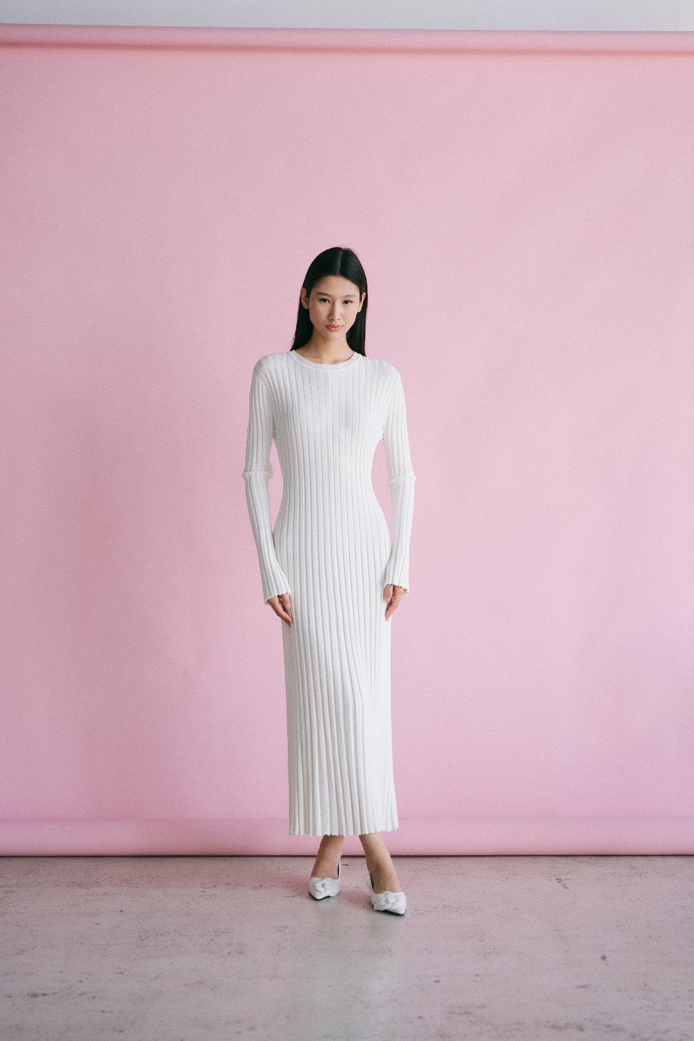 A person with long black hair stands against a light pink backdrop, wearing the Abel Dress that highlights a figure-skimming silhouette and features a pleated texture. The dress, complete with long sleeves and an ankle-length cut, pairs seamlessly with their white shoes as they face the camera directly.