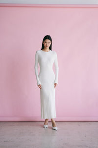 A person with long black hair stands against a light pink backdrop, wearing the Abel Dress that highlights a figure-skimming silhouette and features a pleated texture. The dress, complete with long sleeves and an ankle-length cut, pairs seamlessly with their white shoes as they face the camera directly.