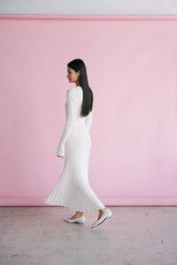 A woman with long dark hair is walking across a pink background in an Abel Dress. She is wearing the Abel Dress, which features long sleeves and a ribbed white fabric that complements her figure-skimming silhouette. Her profile is visible as she walks to the left on a light-colored, speckled surface, paired with white shoes.