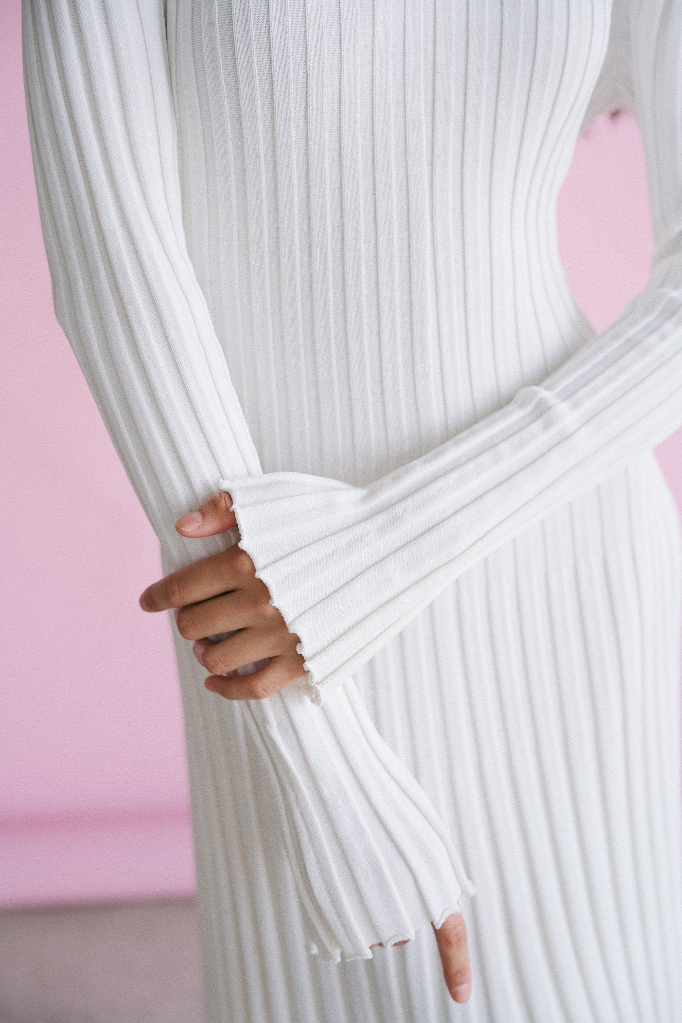 A person wearing a long-sleeved Abel Dress, showcasing its ribbed pleats, stands with their hands gently clasped in front of them. The soft pink background provides a contrasting backdrop, drawing attention to the textured elegance of the dress and the graceful positioning of their hands.