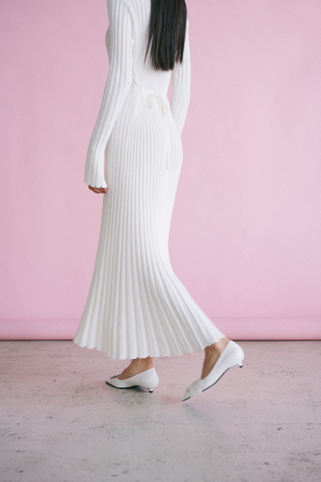 A person with long black hair is standing in a white, pleated-textured Abel Dress with long sleeves, paired with white pointed-toe shoes. The background is pink, and the floor is light grey concrete. Their face is not visible, and they are positioned sideways.