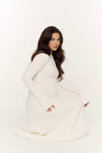 A woman with long, wavy dark hair kneels against a plain white background. She is wearing the Abel Dress, which features long sleeves, a ribbed-knit texture, and a maxi length with pleats. She gazes directly at the camera with a serious expression.