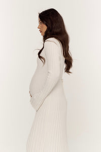 A pregnant woman with long, dark hair, gently cradling her baby bump with both hands, stands in profile against a plain white background while wearing a beige Abel Dress featuring a ribbed and pleated texture.