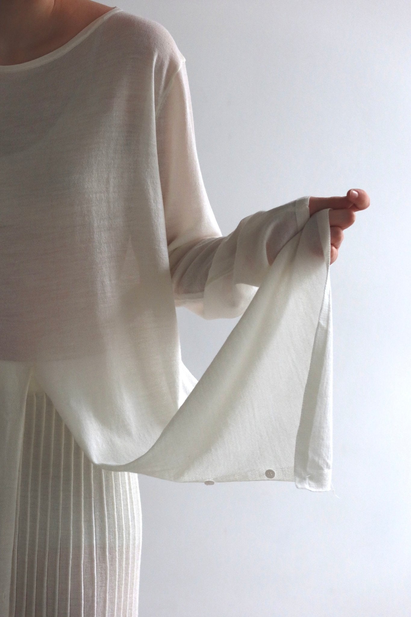 A person wearing a sheer, white, long-sleeved top is holding out the sleeve with their right hand, showcasing the fabric’s light and translucent texture. Paired with the ruffled skirt from the **Alaya Set**, this ensemble exudes elegance against the plain white wall, highlighting its delicate materials.