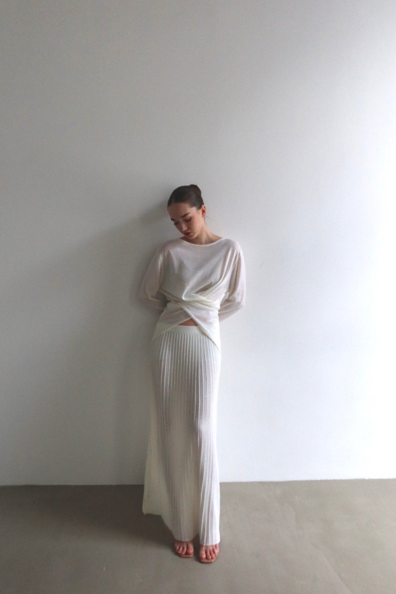 A person stands against a plain white wall, clad in the Alaya Set, which includes a buttersoft long white skirt paired with a matching long-sleeve top. With hands folded in front and head slightly tilted down, they create a serene and minimalist aesthetic.