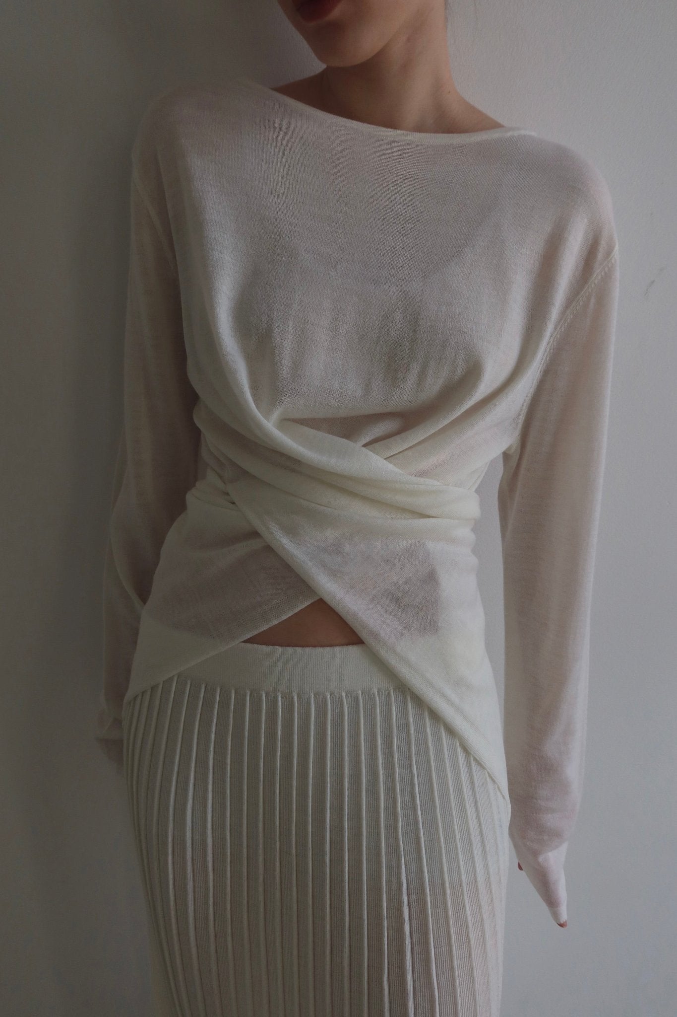 A person dressed in the Alaya Set, which includes a sheer white long-sleeve top with a wrap design paired with a buttersoft pleated skirt, stands against a plain white background. The photo is cropped to only show the clothing, omitting the upper part of their face.