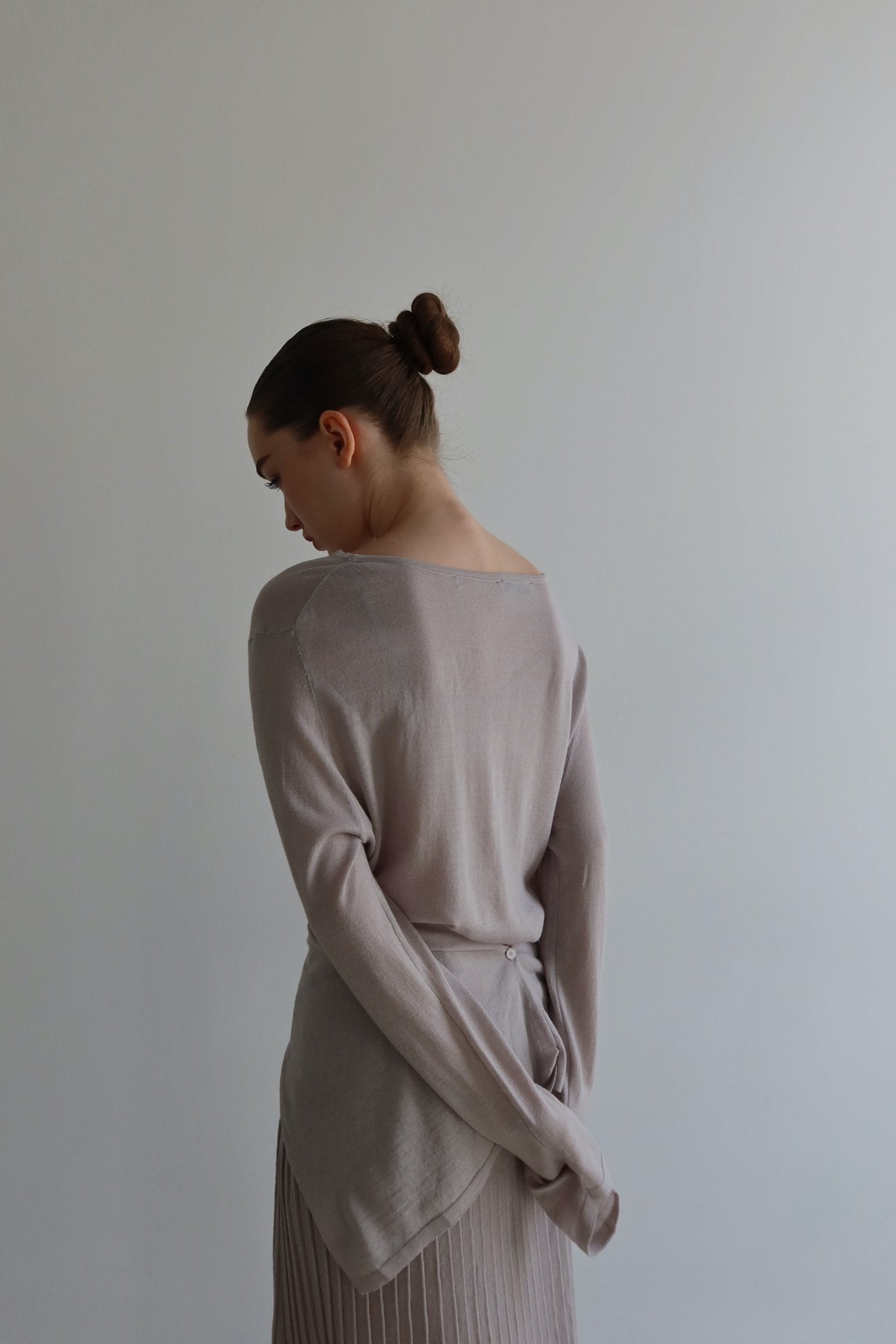 A woman with dark hair in a bun, wearing the Alaya Set, stands with her back to the camera in a minimalist setting. Her light gray top and ruffled skirt, crafted from buttersoft material, feature a button detail at the back hem. With her arms folded behind her back, she is highlighted against a plain white backdrop.