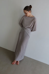 A woman with dark hair tied in a bun is standing against a plain wall. She is wearing the Alaya Set—a light gray, long, pleated skirt and a matching long-sleeve top made of buttersoft material that wraps around her waist. Standing barefoot, she looks down with her hands behind her back.