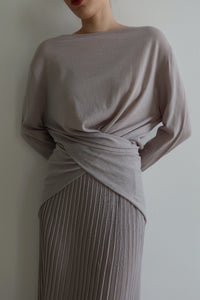 A person is dressed in the Alaya Set, showcasing a long-sleeved, light grey top with an elegant wrap detail at the waist, complemented by a coordinating pleated skirt. The buttersoft material flows gracefully as they pose against a plain white background, their head gently tilted.