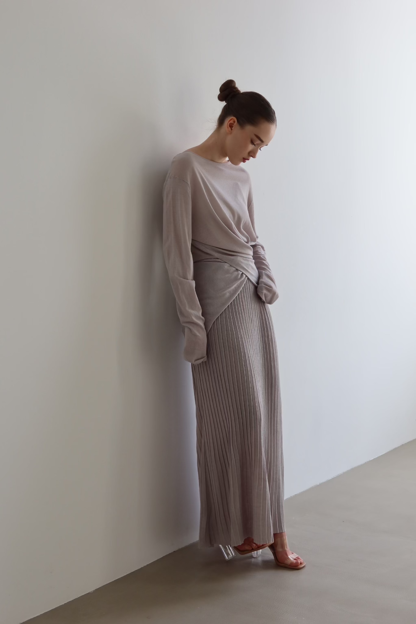 A person with their hair tied back in a bun leans against a white wall, looking down. They're wearing the Alaya Set, which includes a long-sleeved, light grey top made of buttersoft material paired with a matching pleated maxi skirt. With one hand in a pocket and light-colored open-toe sandals, they exude effortless elegance.