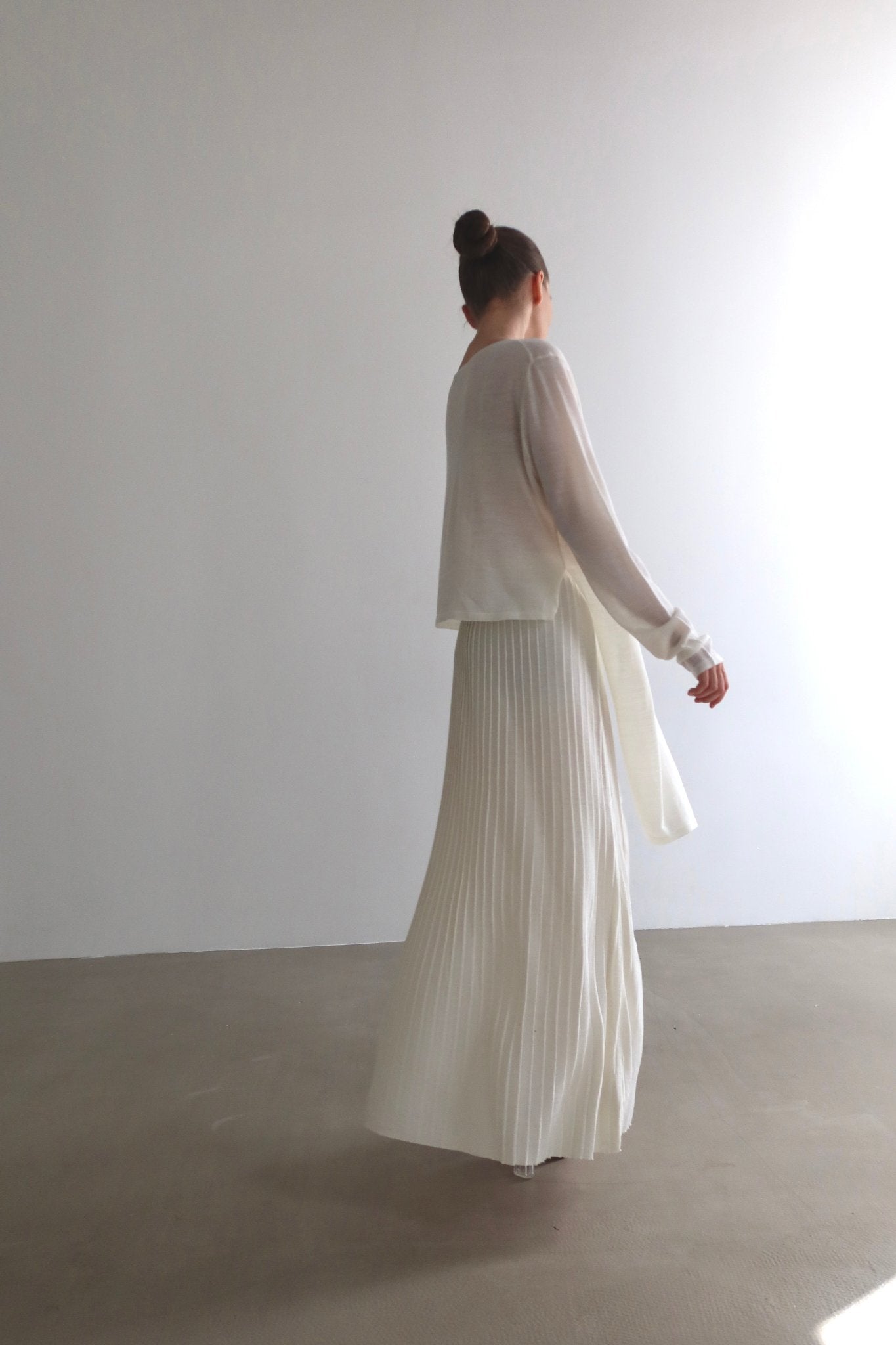 A person with their hair in a bun stands with their back to the camera, showcasing a light-colored, long-sleeved top and a matching ruffled skirt from the Alaya Set. The backdrop features a minimalist interior with white walls and a smooth, neutral-toned floor.